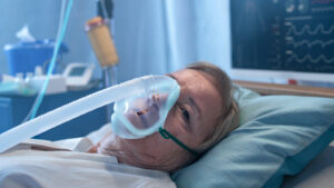 A sick woman lies in a hospital bed. She is wearing an oxygen mask.